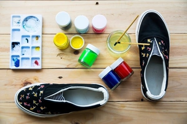 Пара черных холщовых туфель с ручной росписью на столе с различными красками и кистями.