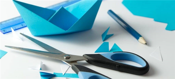 Ножницы и карандаш лежат рядом с синей лодкой оригами.