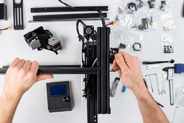 Мужчина в процессе сборки 3D-принтера в окружении различных деталей и инструментов.