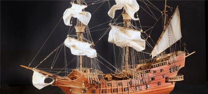 Деревянная модель корабля с мачтами, парусами и такелажем на черном фоне.