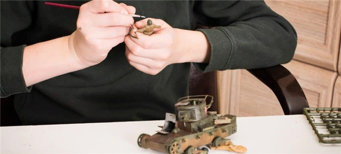 Молодой человек раскрашивает модель солдата с частично собранным танком на столе.