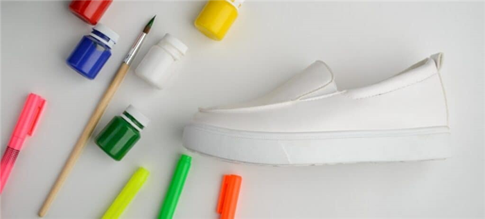 Белая обувь с несколькими яркими банками краски и маркерами для ткани на белом фоне.