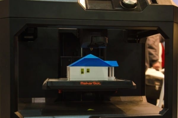 3D-принтер MakerBot Replicator+ заканчивает печать модели дома с синей крышей.