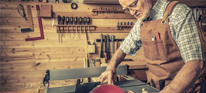 Пожилой мужчина в своей деревообрабатывающей мастерской использует пилу, надевая рабочий фартук.