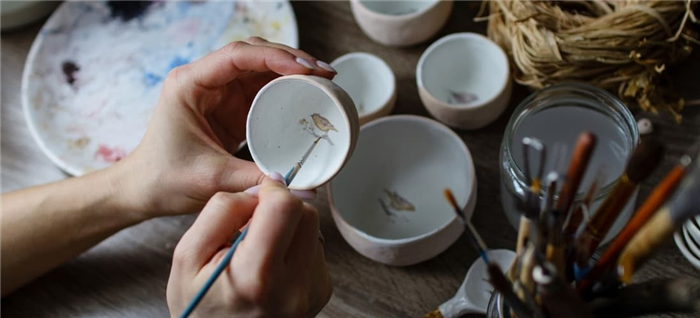 Женщина вручную наносит рисунок птицы на маленькую керамическую чашку.