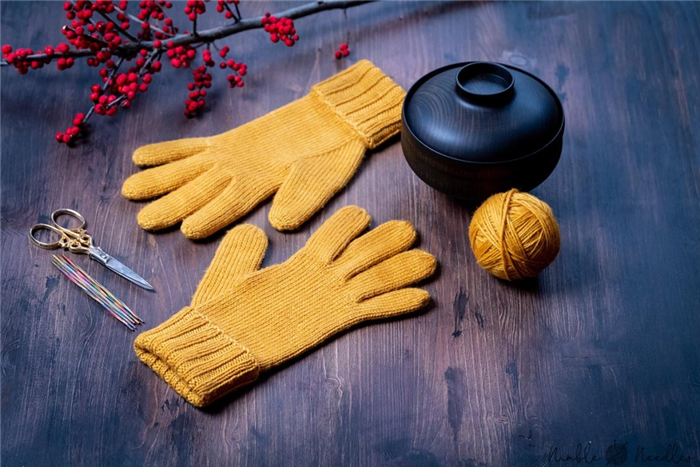 пара желтых вязаных перчаток с пальцами на деревянной доске с инструментами для вязания на заднем плане